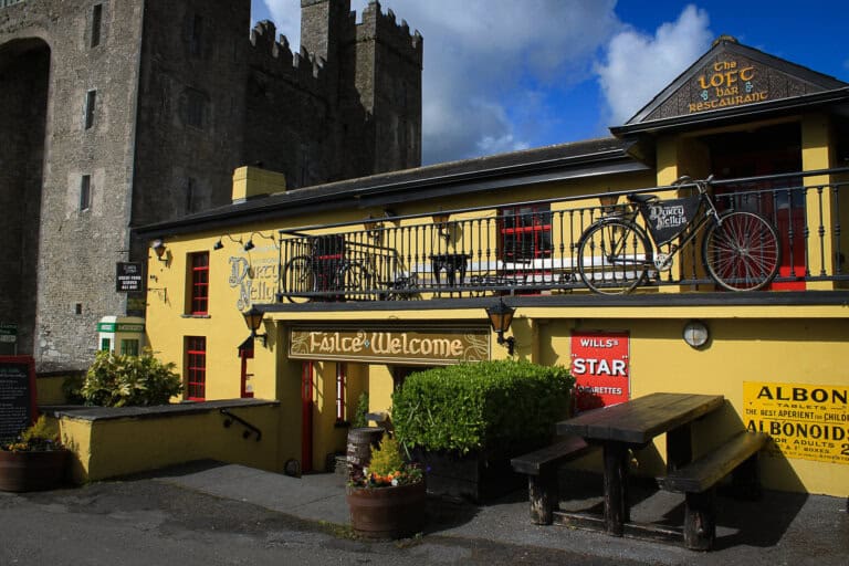 The photo features a vibrant yellow traditional Irish pub named "Dirty Nelly's" with a dark grey roof. Above the entrance, the sign "Fáilte Welcome" greets visitors, indicating hospitality in both Irish and English. A vintage black bicycle is perched decoratively on a balcony above the main door, alongside signage for "The Loft Bar & Restaurant." The pub is adorned with various signs and greenery, adding to its charming character. In the foreground, a rustic wooden bench invites patrons to sit outside. Towering in the background is the grey stone facade of a castle, providing a historic and dramatic backdrop to this quaint establishment. The sky is partly cloudy, suggesting a typical Irish day.