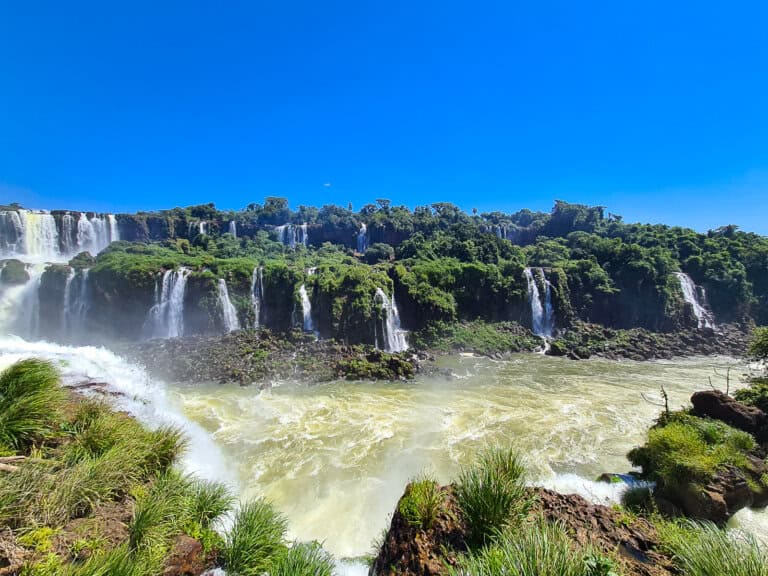 A panoramic view of Iguazú Falls surrounded by lush greenery under a clear blue sky. Multiple cascades of water plummet over the cliff edges, creating a dramatic and powerful natural spectacle.