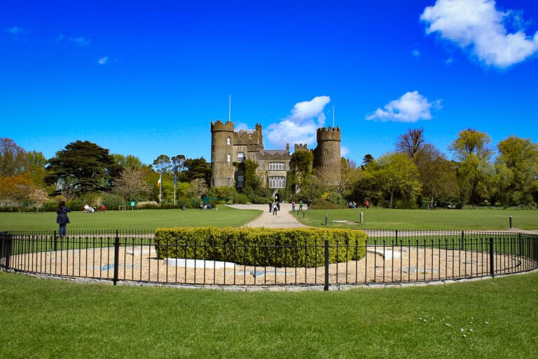 The image features the imposing Malahide Castle in Ireland, a historic structure with grey stone towers and battlements set against a vivid blue sky with fluffy white clouds. The castle is situated amidst a lush green park with well-maintained lawns, trees, and gardens. In the foreground, a circular hedge encloses a sand-filled area, bordered by a black iron fence. Visitors can be seen enjoying the park, with some walking on the central path leading to the castle's entrance. The scene is lively and picturesque, capturing the essence of a sunny day at this heritage site.