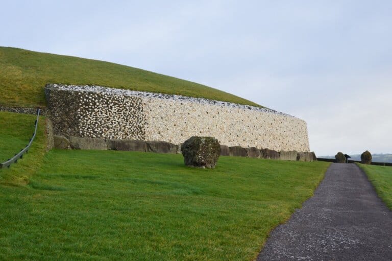 The photo captures the ancient structure of Newgrange in Ireland, a prehistoric monument dating back to around 3200 BC. The image shows a section of the structure's exterior with its unique white quartz facade, resting atop a retaining wall of rustic grey stones. A lush green mound, indicative of the structure's earth-covered tomb, rises above the stone wall. In the foreground, a well-defined gravel path invites visitors to approach, flanked by verdant grass and occasional standing stones that add to the site's mystique. The sky is overcast with a break in the clouds allowing a soft light to enhance the scene's calm and historical ambiance.