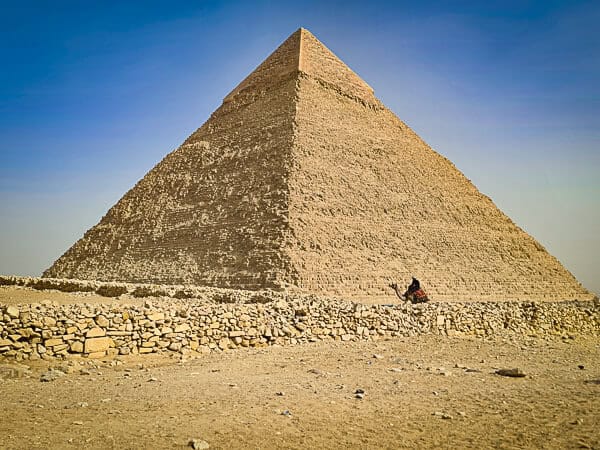 The image showcases one of the iconic Great Pyramids of Giza in Egypt, with its immense triangular façade composed of large limestone blocks. There's a clear blue sky above, and in the foreground, a person on horseback appears diminutive in comparison, emphasizing the pyramid's monumental scale. The ground is arid and strewn with stones and rubble, indicative of the desert environment surrounding the pyramids. The angle of the shot captures the pyramid's precise geometric shape and the craftsmanship that has stood the test of millennia.