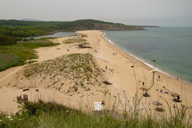 The image provides an elevated view of Veleka Beach near Sinemorets, a picturesque spot where the Veleka River meets the Black Sea. The beach forms a unique natural landscape with its riverbank on one side and the sea on the other, separated by a strip of golden sand. Visitors can be seen enjoying the sun and the relatively uncrowded beach, suggesting a peaceful and natural setting away from the bustle of more popular tourist destinations. A wooden observation deck overlooks the river, offering a spot to take in the scenic view. The backdrop of lush green hills indicates a rich, biodiverse area, possibly part of a protected nature reserve. The overcast sky suggests a mellow atmosphere, with conditions that are comfortable for spending time outdoors without the harshness of direct sunlight. The image beautifully captures the tranquility and unspoiled beauty of this coastal area.