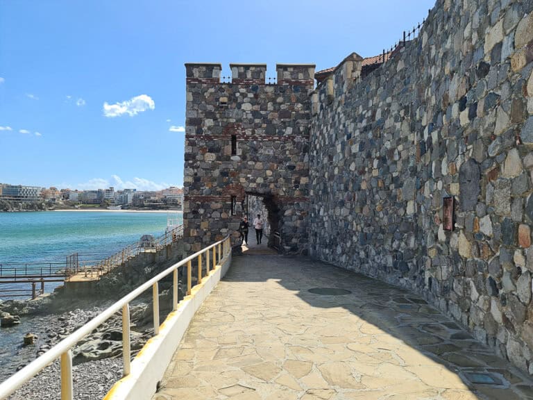 The image depicts a fortified stone walkway leading to a robust gatehouse by the sea. The walkway is lined with a metal railing on the side facing the water, ensuring the safety of pedestrians. The stone construction of the walls and gatehouse suggests historical or architectural significance, possibly part of an old fortress or city walls. The wall is composed of varied shapes and colors of stone, adding texture and visual interest. Beyond the gatehouse, a calm sea extends to the horizon, with a view of a modern cityscape across the water. The buildings across the sea are contemporary, with what appears to be residential and possibly commercial structures. Clear skies with a few clouds indicate a pleasant day, perfect for a walk along this historic path. People can be seen walking through the gatehouse, hinting at a popular route for tourists and locals alike, merging the past with present-day leisure activities.