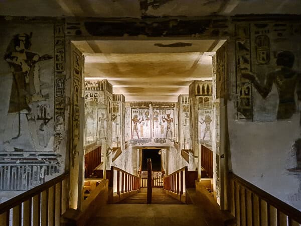 This image captures the interior of an ancient Egyptian tomb or temple passageway. The walls are adorned with well-preserved hieroglyphs and frescoes, showing figures and symbols that were significant in the context of Egyptian mythology and funeral rites. The corridor descends towards the inner sanctum, with wooden railings leading down a staircase to guide visitors. The lighting is subtle, highlighting the colors and details of the wall carvings, and the ceiling appears to have remnants of decorative elements as well. This passageway serves as both a historical archive and a path deeper into the heart of the structure, where sacred or burial chambers might be found.