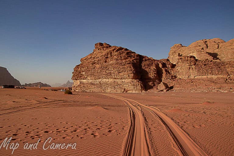 Scenic view of the Wadi Rum desert in Jordan, featuring red sand dunes, rugged rock formations, and vehicle tracks under a clear blue sky, a must-see destination included in any Jordan 5-day itinerary.