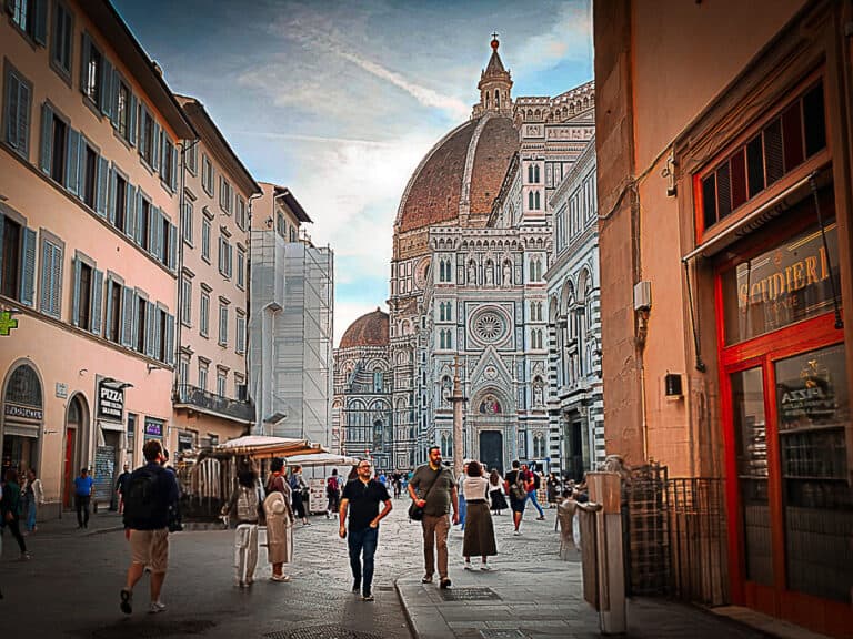 Pedestrians walk along a bustling street in Florence, Italy, with a stunning view of the Florence Cathedral (Duomo) and its iconic dome in the background. Historic buildings and storefronts line the street, capturing the vibrant atmosphere of the city center.