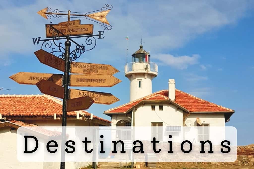 Vintage signpost with multiple wooden direction signs pointing to local attractions near a charming white lighthouse with a red-tiled roof, under a bright blue sky, symbolizing global travel destinations.