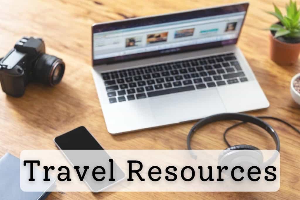 A workspace with a laptop displaying a travel planning website, surrounded by a camera, headphones, smartphone, and a potted plant, representing essential travel resources and tools.
