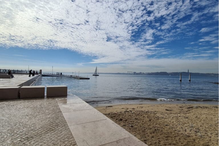 A serene Tagus River waterfront view in Lisbon, featuring sailboats, a boardwalk, and blue skies. Hotels near the river provide a peaceful escape while still being close to the city's historic center—perfect for travelers searching for where to stay in Lisbon with river views.