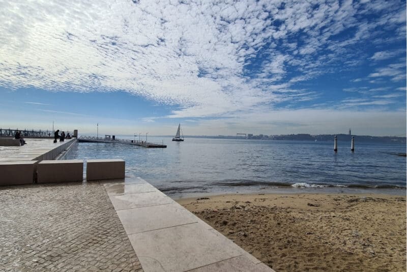 A serene Tagus River waterfront view in Lisbon, featuring sailboats, a boardwalk, and blue skies. Hotels near the river provide a peaceful escape while still being close to the city's historic center—perfect for travelers searching for where to stay in Lisbon with river views.