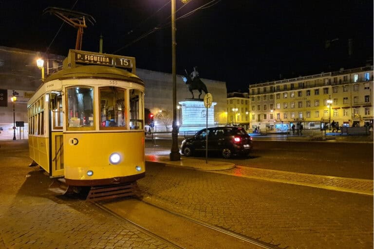 A classic yellow tram in Lisbon, stopped at Figueira Square (Praça da Figueira) at night, with the illuminated statue of King João I in the background. One of the best Lisbon travel tips is to ride the famous tram 28 for a scenic city tour.