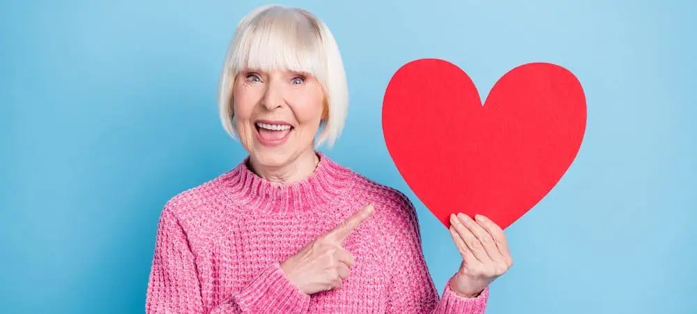 A smiling woman holding up and pointing to a cardboard cut-out of a heart