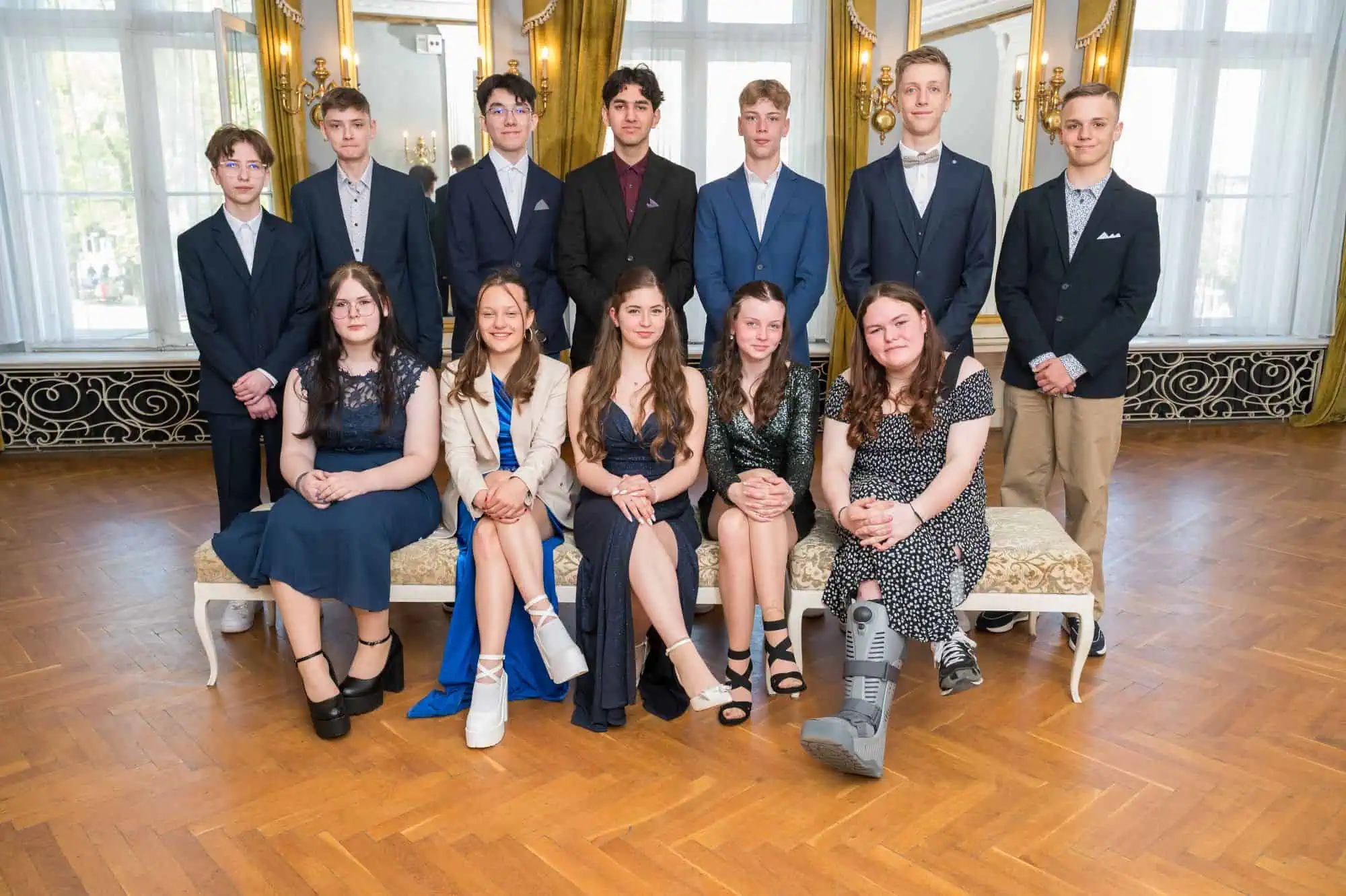 Gruppenfoto von Jugendlichen bei der Jugendweihe im Barocksaal Rostock, elegant gekleidet, stehend und sitzend in prunkvoller Kulisse mit goldenen Spiegeln und Parkettboden.