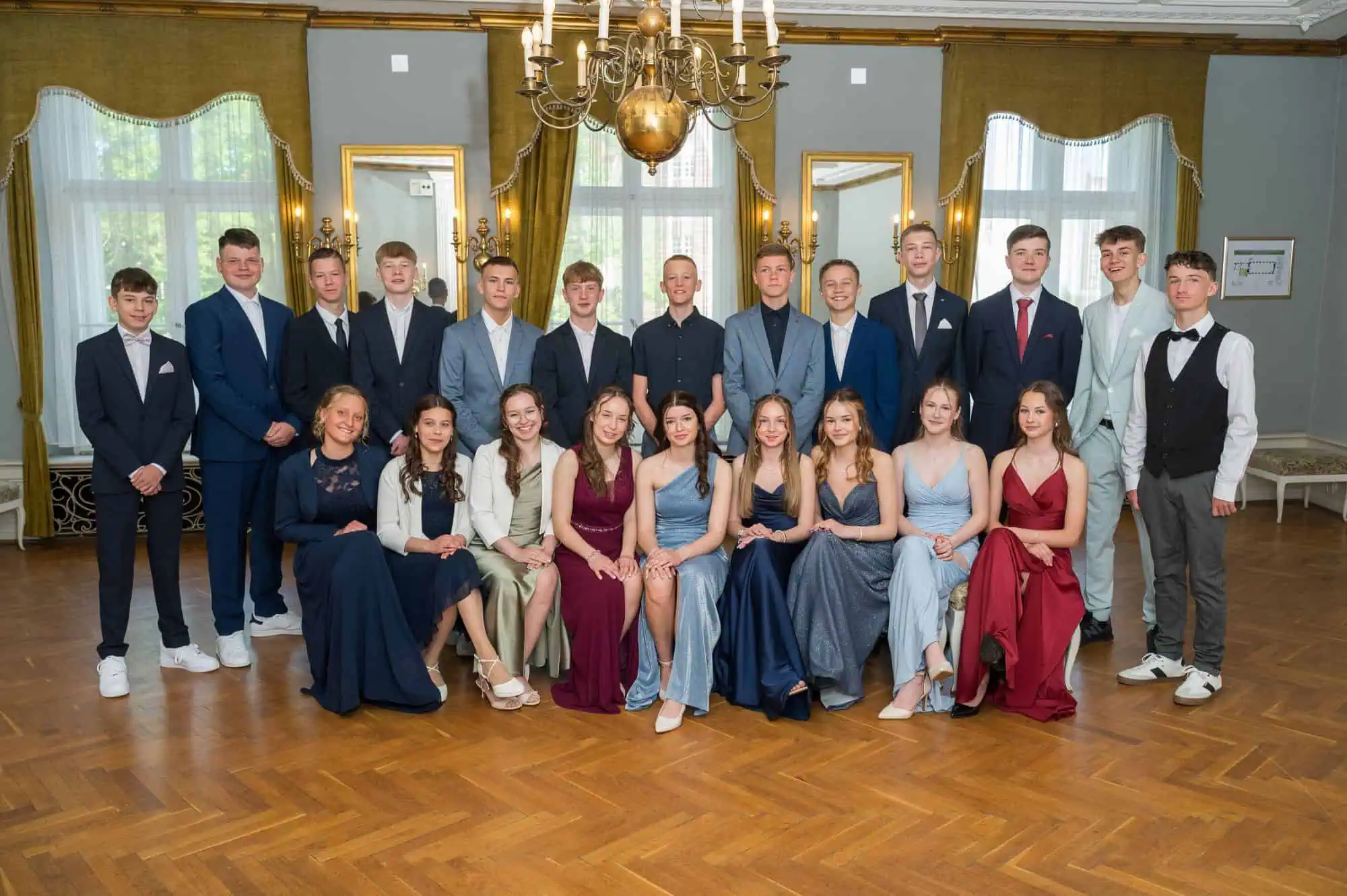 Jugendweihegruppe am 03.05.2025 im Rostocker Barocksaal, aufgenommen vom Rostocker Fotografen, junge Frauen in eleganten Kleidern und junge Männer in Anzügen, vor prunkvoller Kulisse mit Kronleuchter.