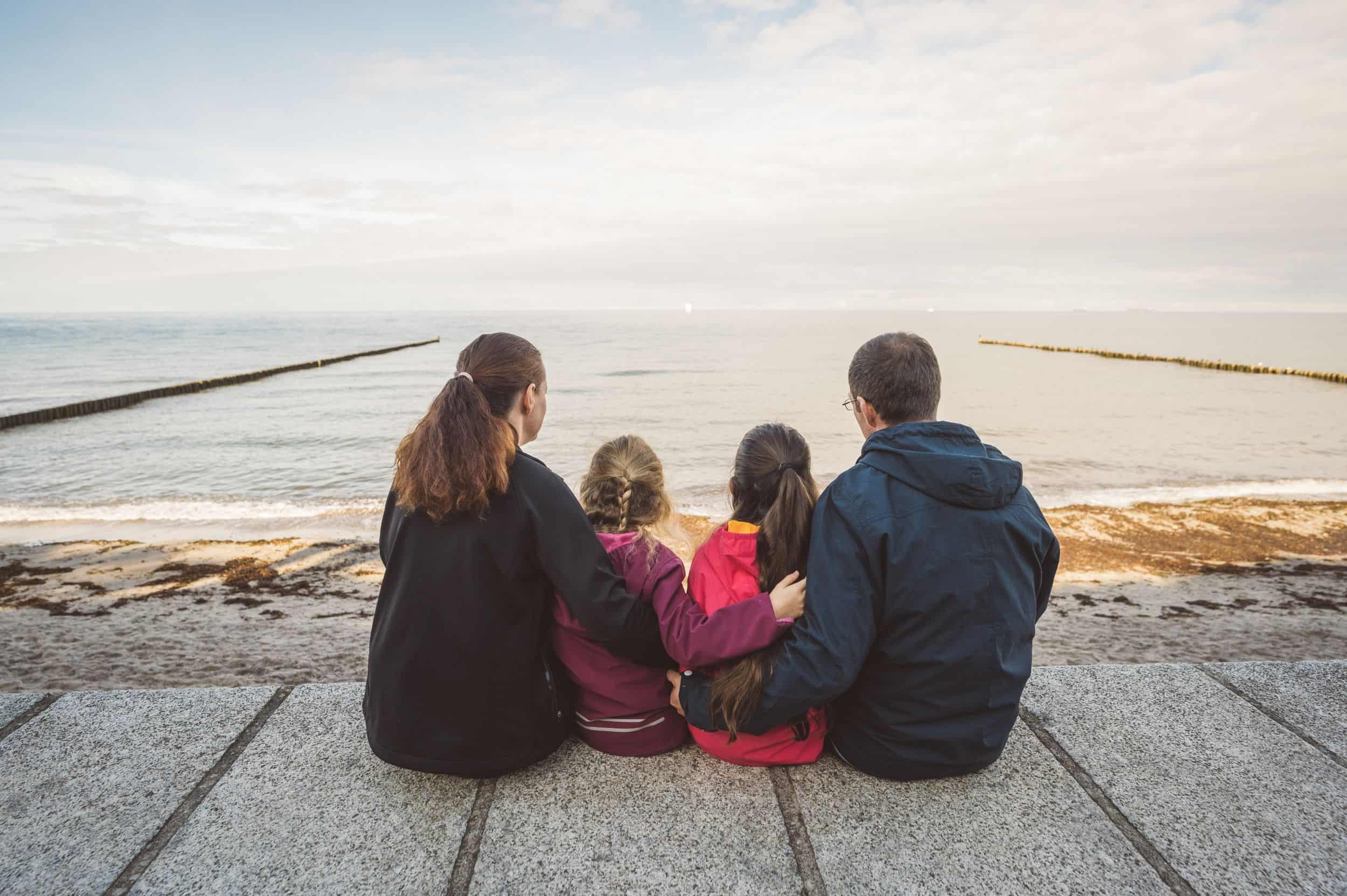 Familienfoto am Strand von Nienhagen beim Gespensterwald Familie mit zwei Kindern sitzt Rücken an Rücken auf einer Promenade am Meer und blickt gemeinsam in die Ferne über das Wasser.