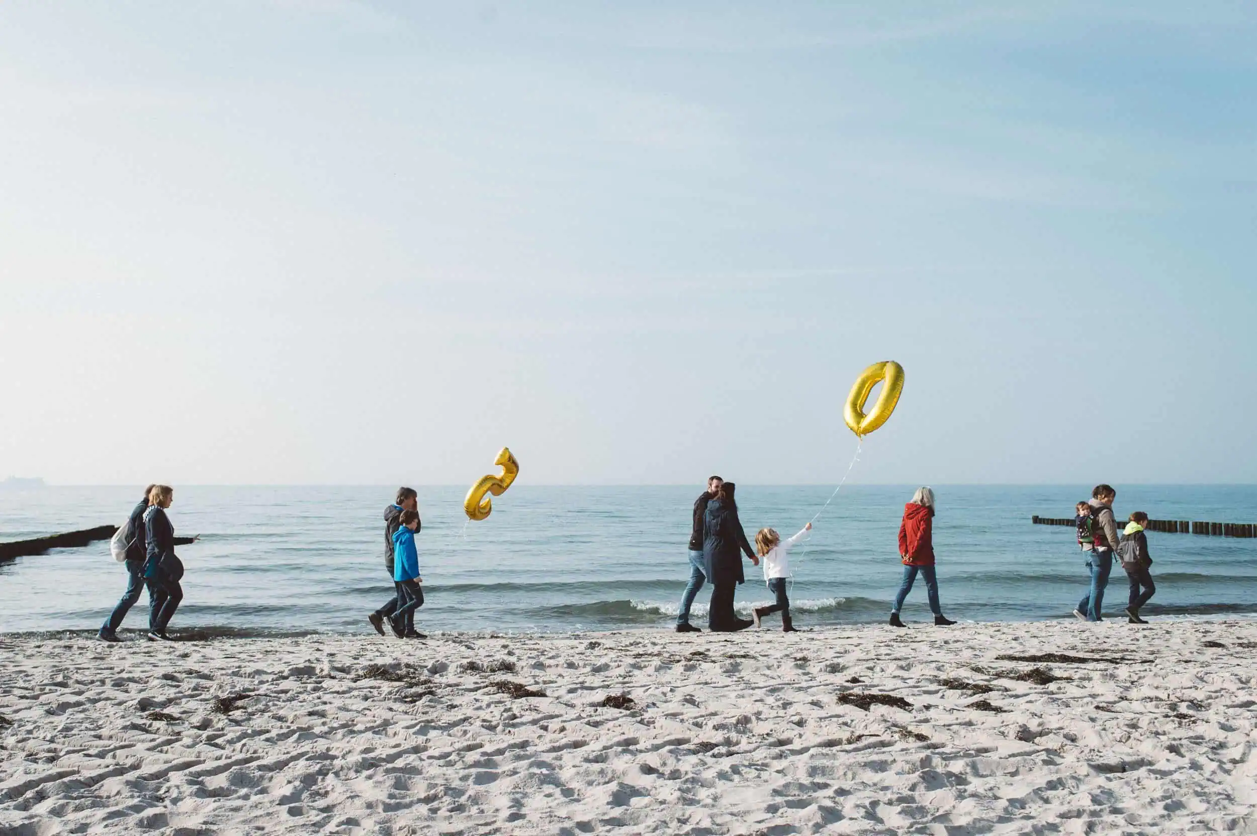 Natürliche Familienfotos am Strand – echte Momente mit Meerblick Mehrere Familien spazieren mit Kindern und goldenen Luftballons in Form von Zahlen am Strand entlang – natürliche Familienfotos mit Ostsee-Flair