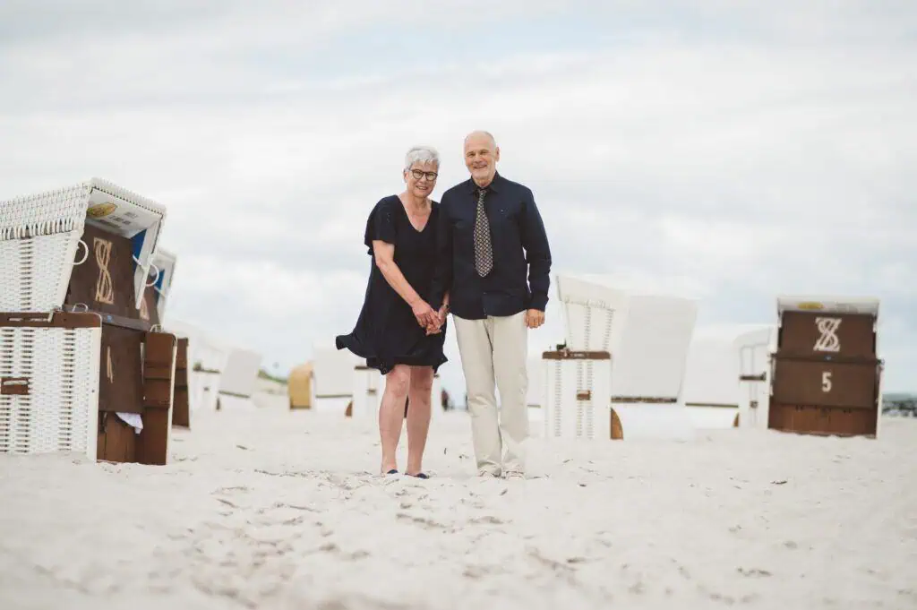 Älteres Paar hält Händchen am Sandstrand von Ahrenshoop vor Strandkörben und lächelt glücklich in die Kamera – harmonischer Moment an der Ostsee unter bewölktem Himmel.