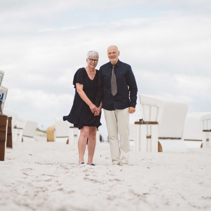 Verliebtes Seniorenpaar am Strand von Ahrenshoop – Lebensfreude an der Ostsee Älteres Paar hält Händchen am Sandstrand von Ahrenshoop vor Strandkörben und lächelt glücklich in die Kamera – harmonischer Moment an der Ostsee unter bewölktem Himmel.