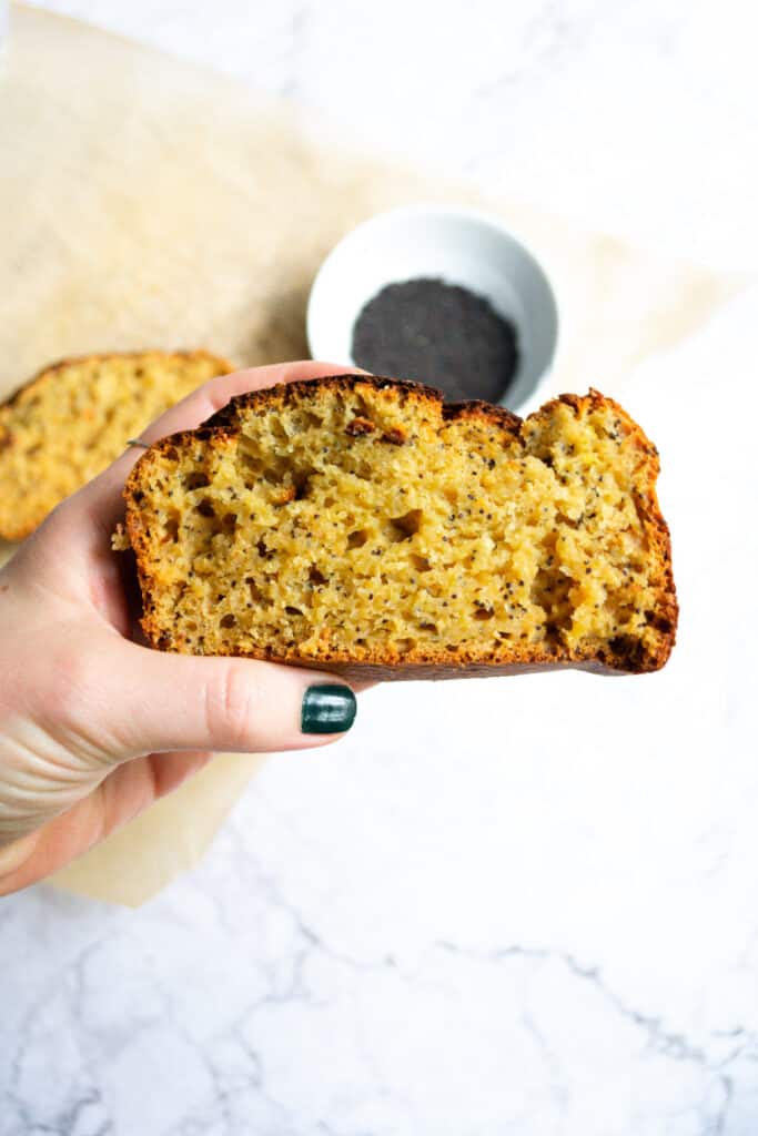 Hand holding a slice of orange poppyseed loaf with poppyseeds in the background.