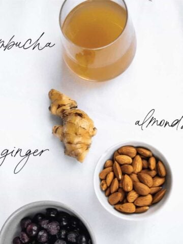 An overhead shot showing a glass of kombucha, a knob of ginger, and a bowl of almonds on a white background.