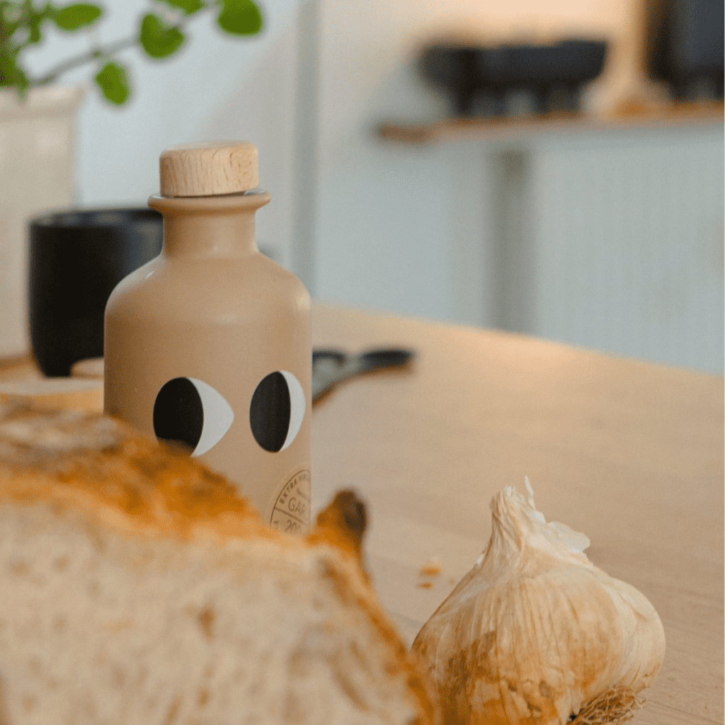 Bottle of Yiayia Extra Virgin Olive Oil with Garlic being cheeky and hiding behind a loaf of bread.