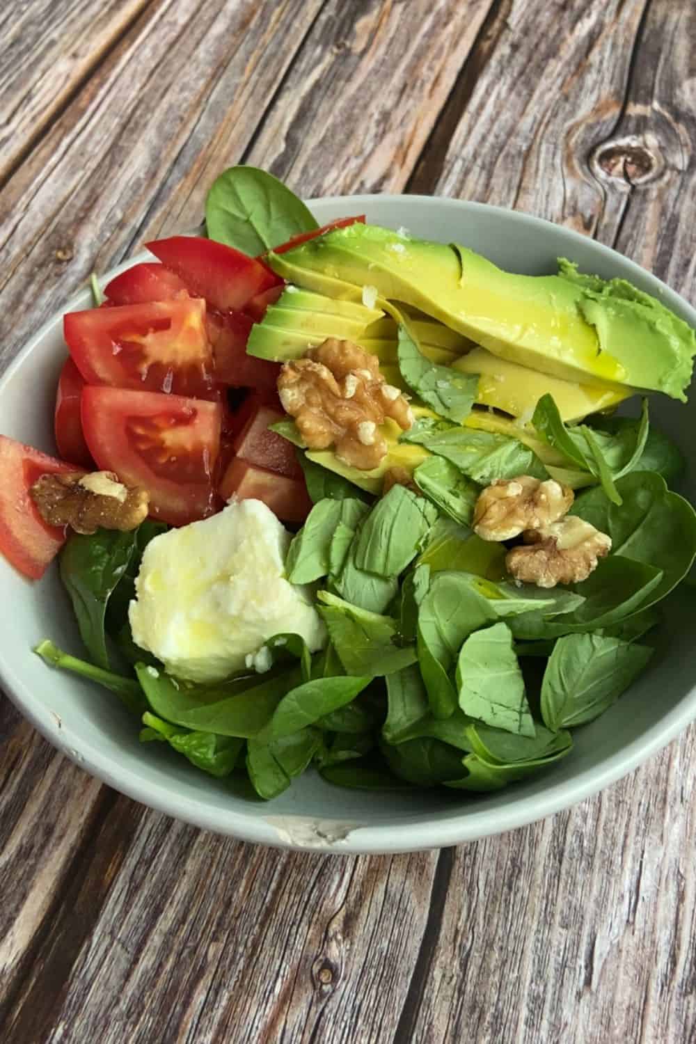 A photo of a spinach, avocado and tomato salad in a bowl on a wooden table with walnuts on top.