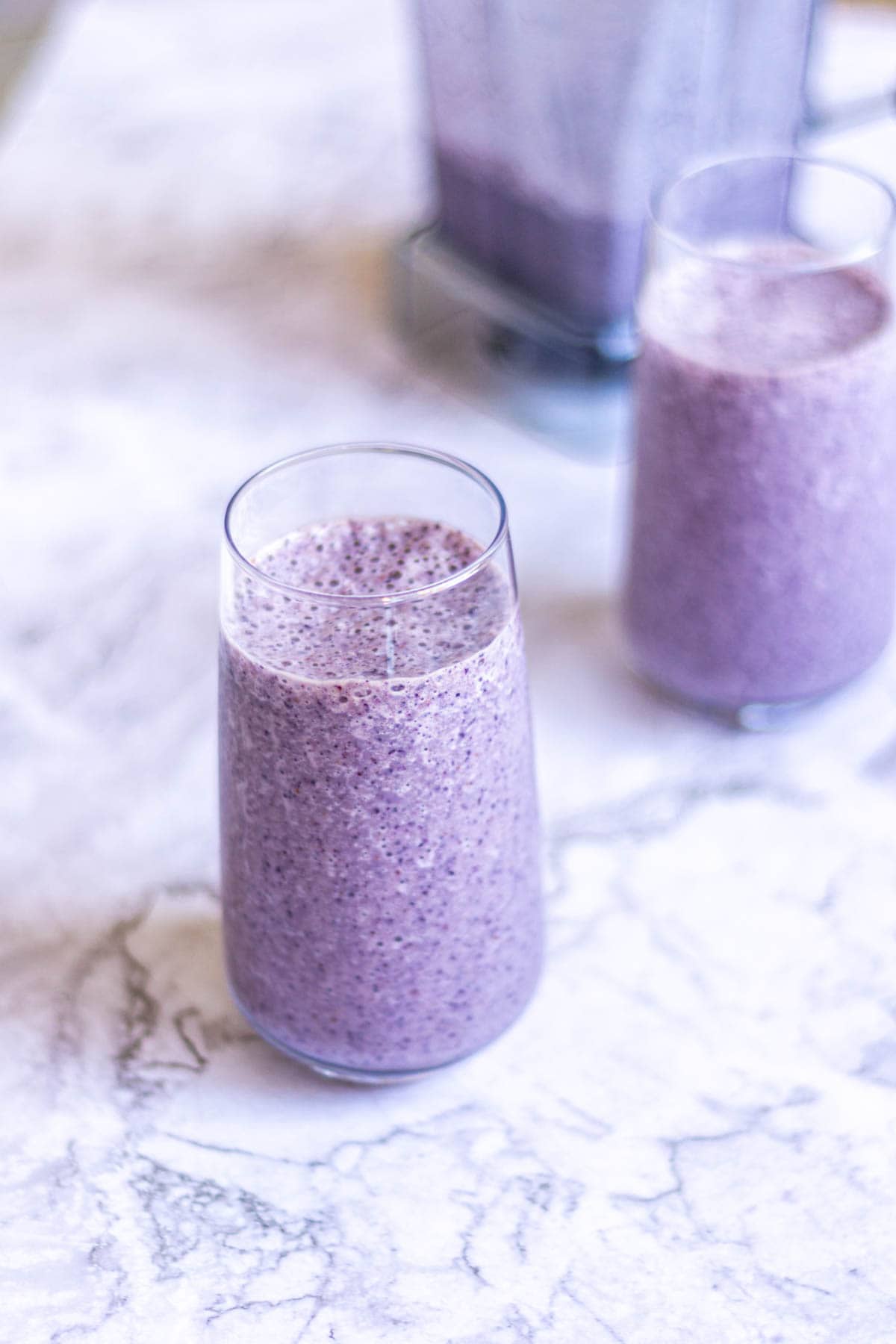 Two glasses of a purple smoothie, with a blender in the background blurred out.