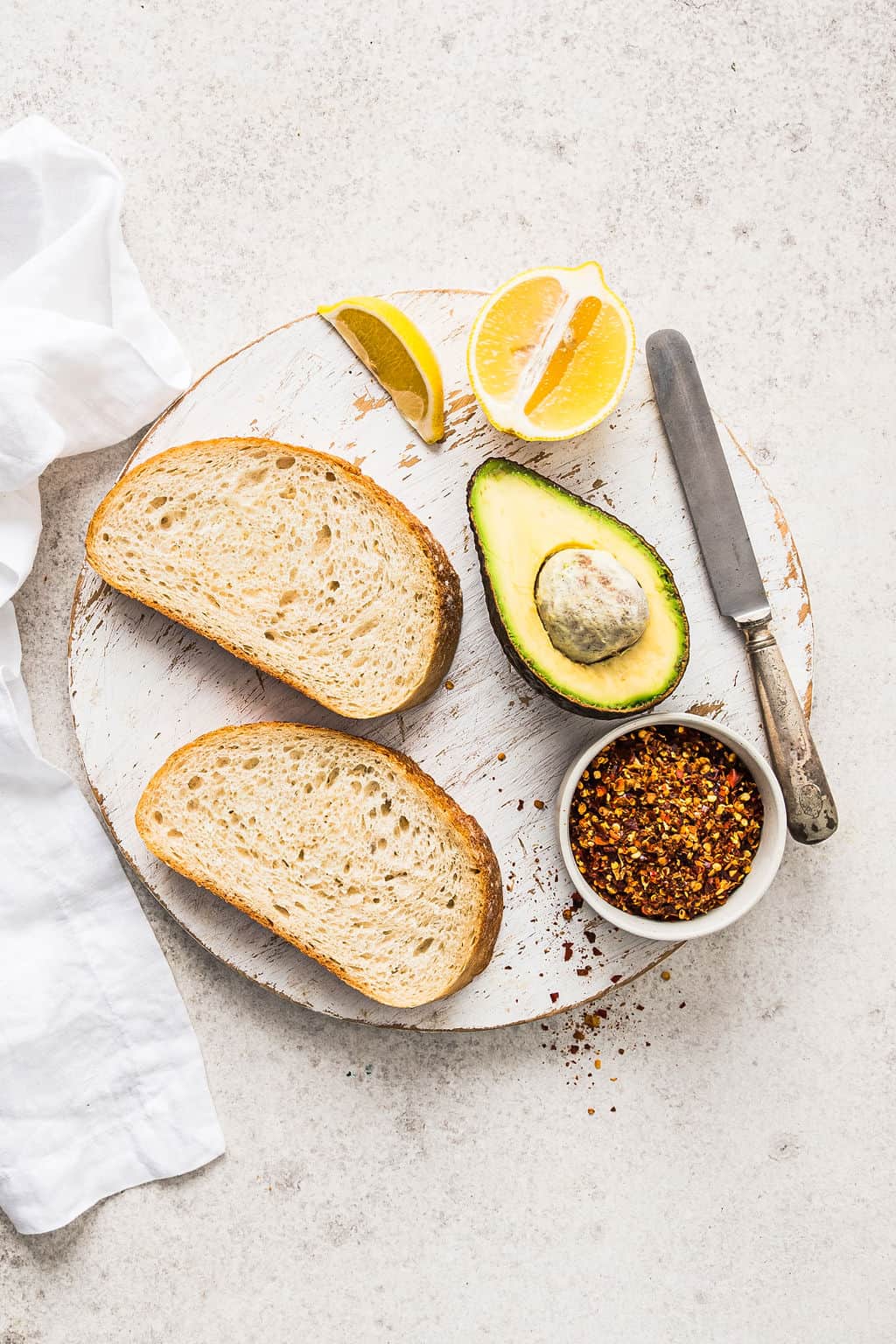 Overhead shot of basic avocado toast ingredients including bread, avocado, lemon and chilli flakes.