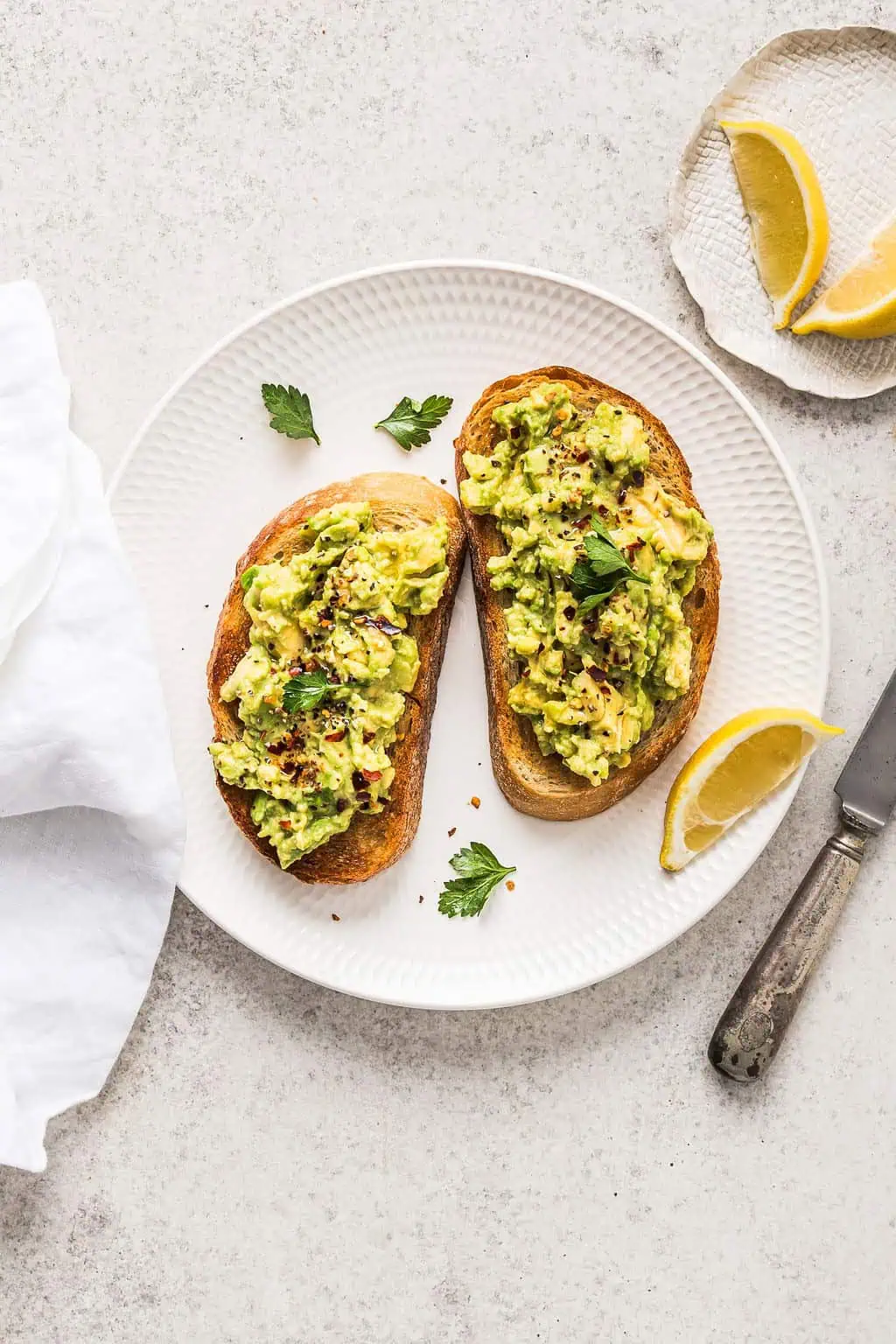 Overhead shot of basic avocado toast on a plate, topped with parsley, lemon and chilli flakes.
