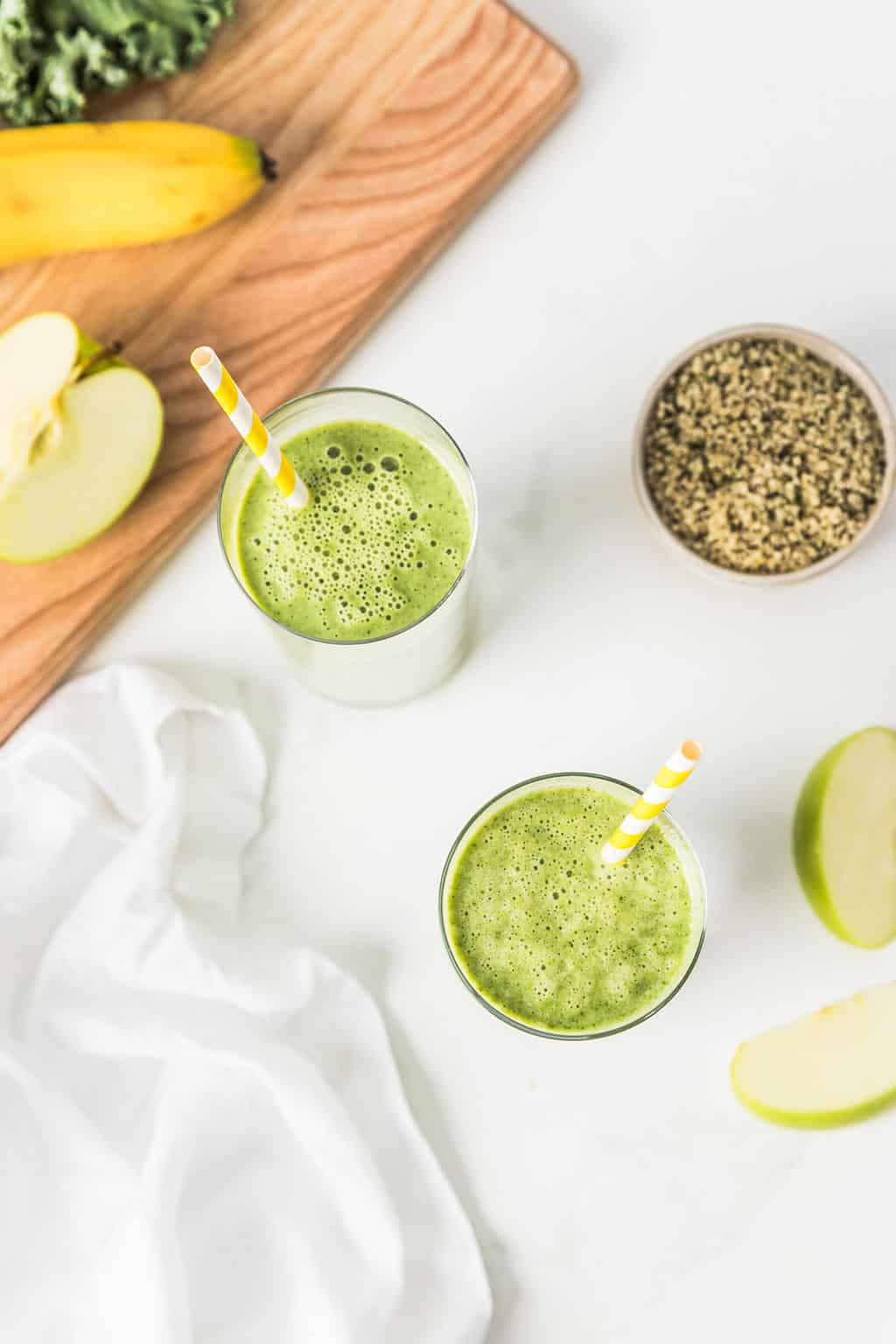 Overhead shot of two kale smoothies in glasses, you can also see hemp seeds, apples and bananas in the photo.