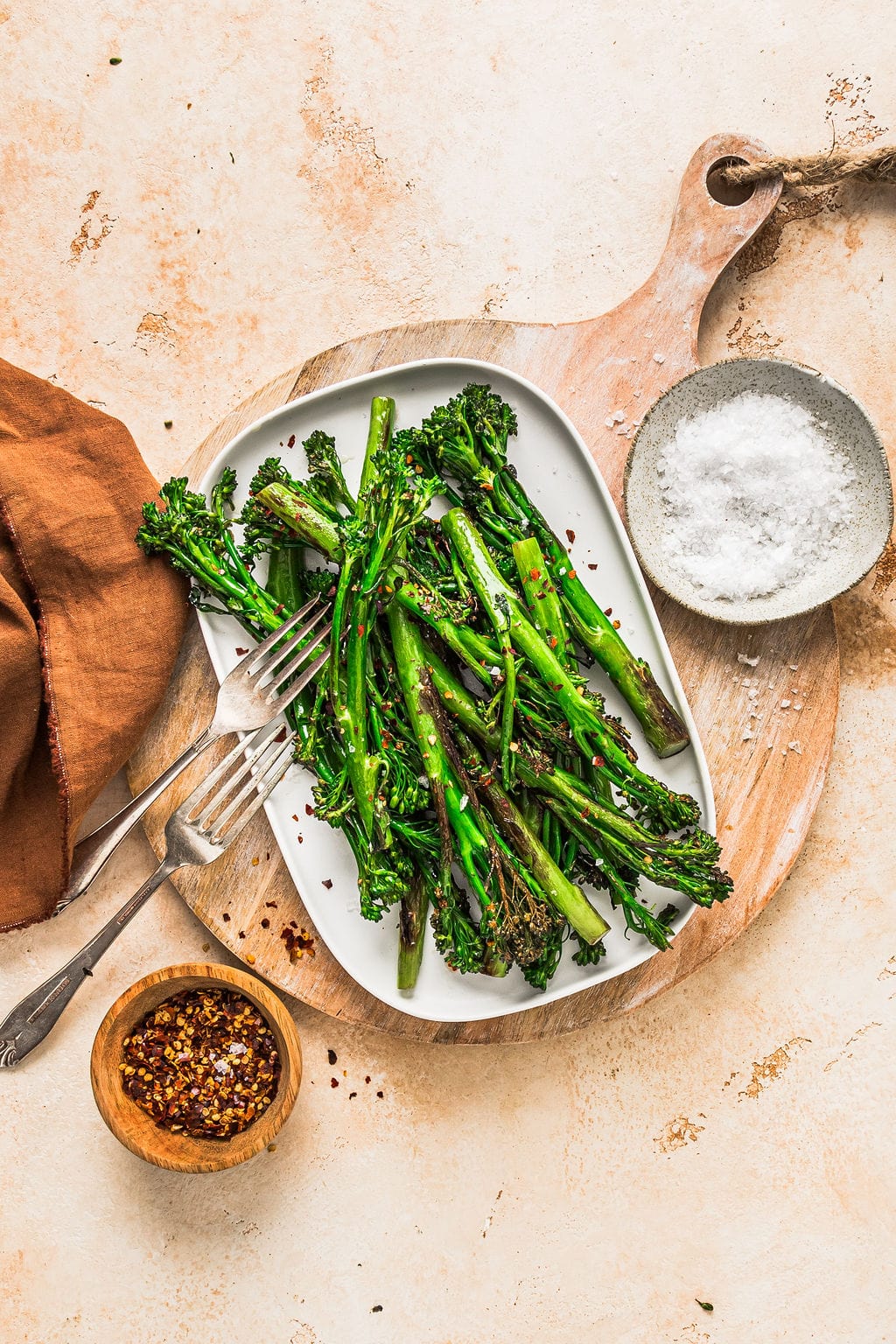 Overhead view of pan fried broccolini on a white plate, on top of a tan coloured table.
