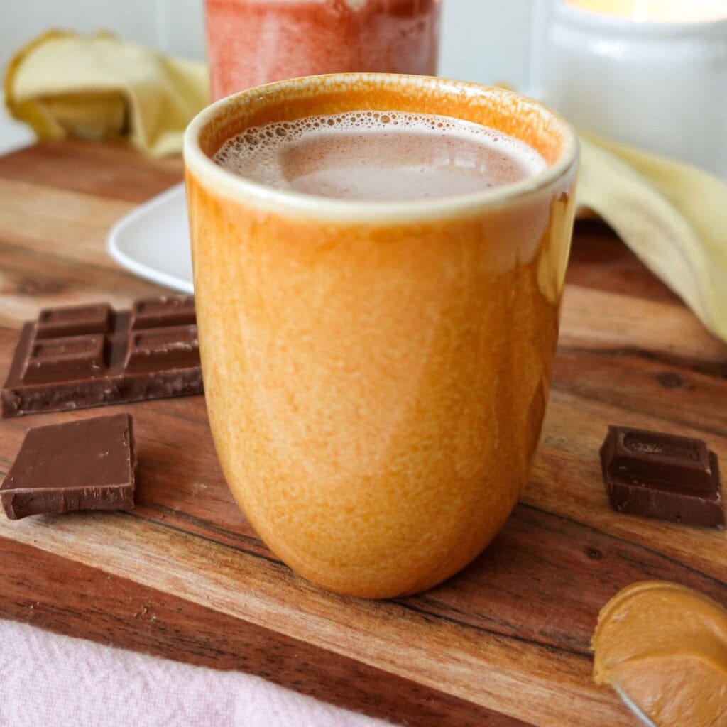 A closeup of Biscoff hot chocolate in an orange mug with a spoon of Biscoff spread and chocolate pieces in the foreground.