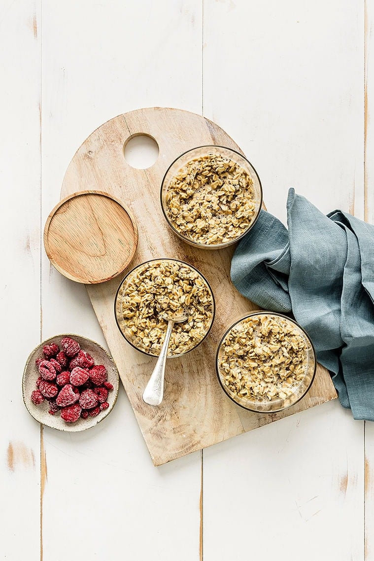 Overhead shot of 3 jars of meal prep overnight oats on a cutting board, next to a bowl of raspberries.