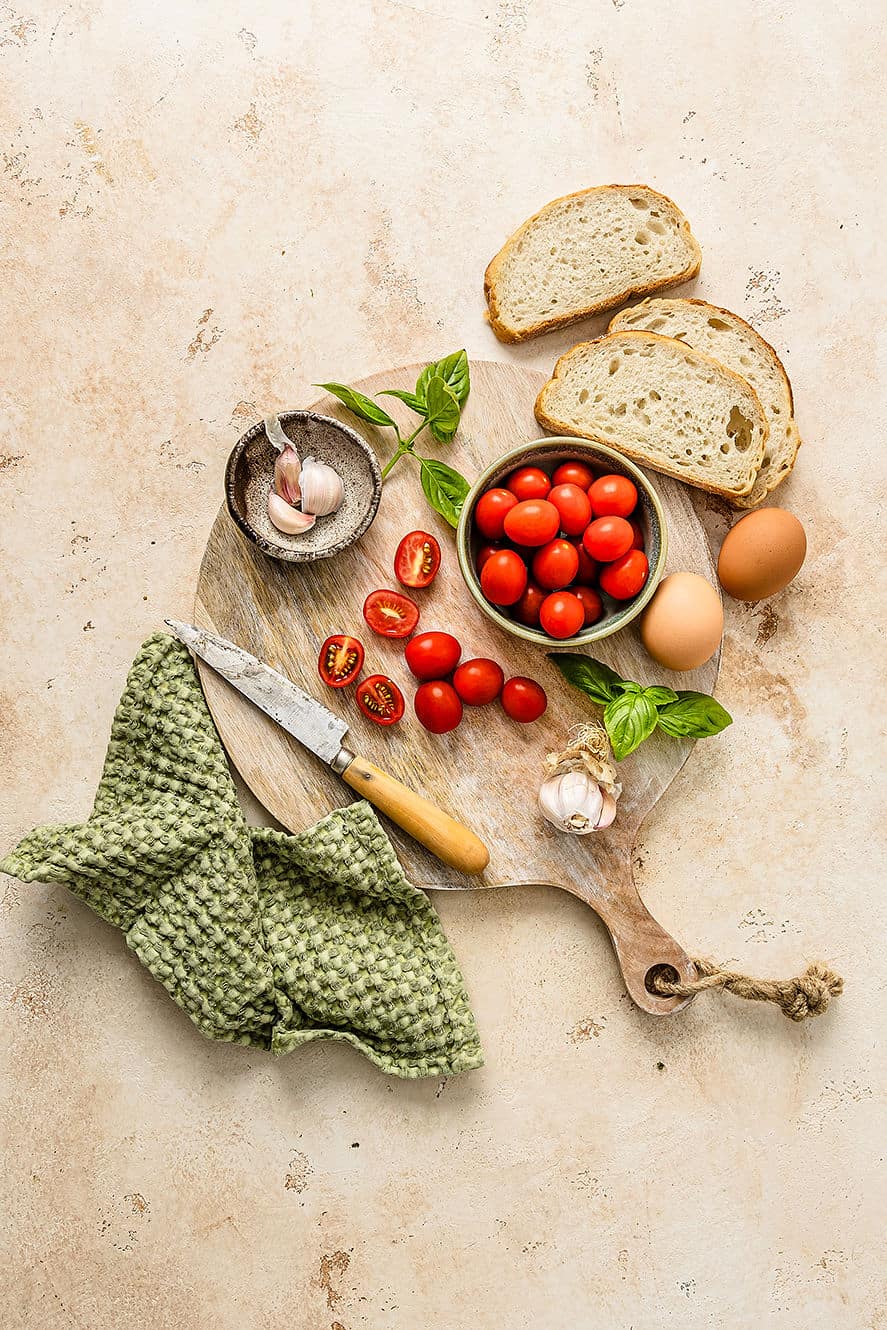 Overhead shot of breakfast bruschetta ingredients including sourdough bread, cherry tomatoes, garlic, eggs and basil.