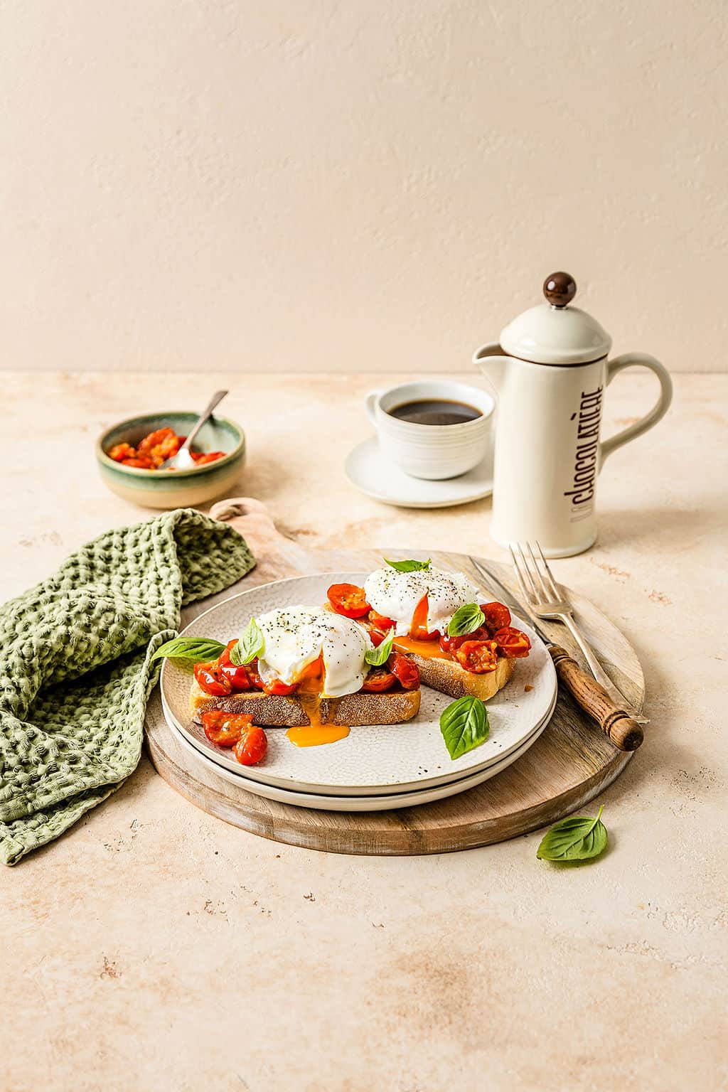 Breakfast bruschetta on a plate, topped with a runny poached egg with a carafe of coffee in the background.