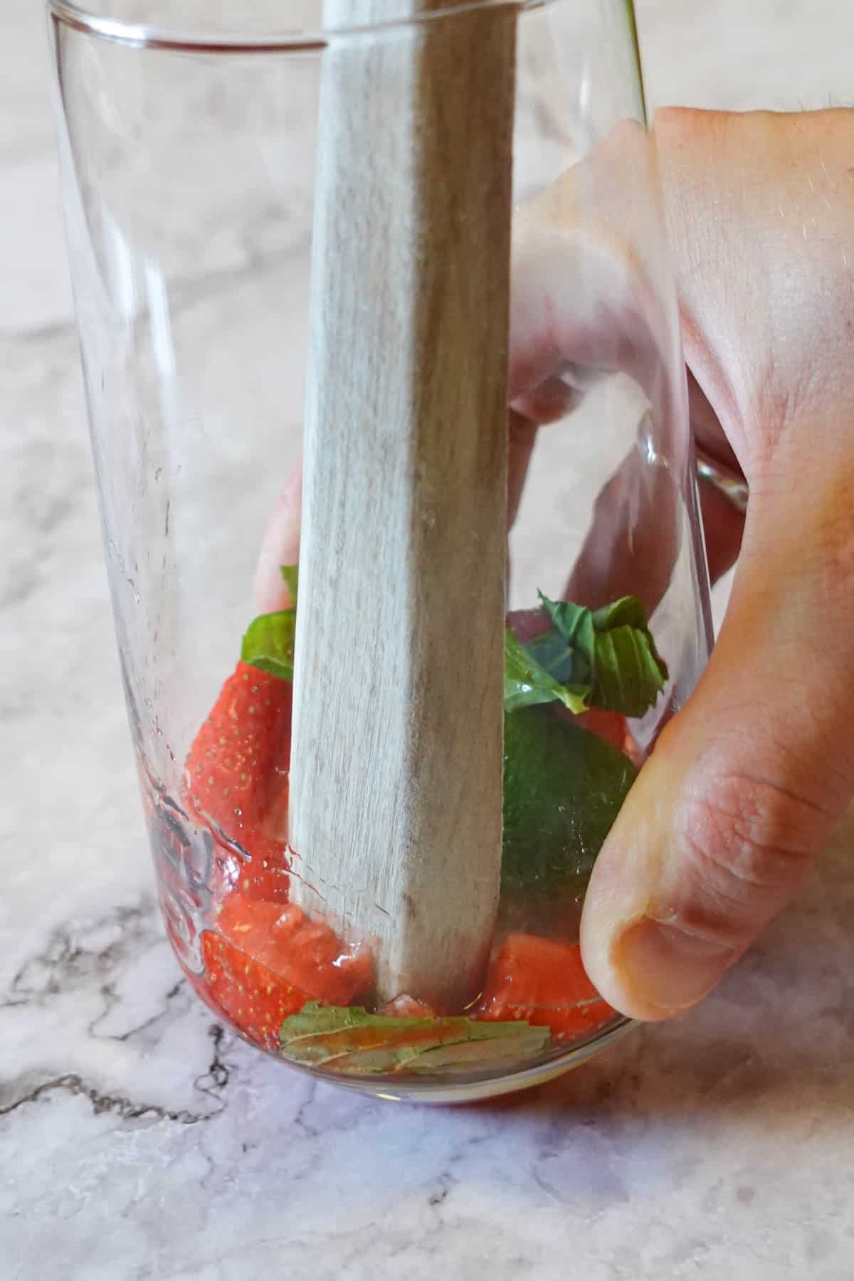 Close up of strawberries, lime wedge, mint leaf and simple syrup being muddled in the bottom of a glass.