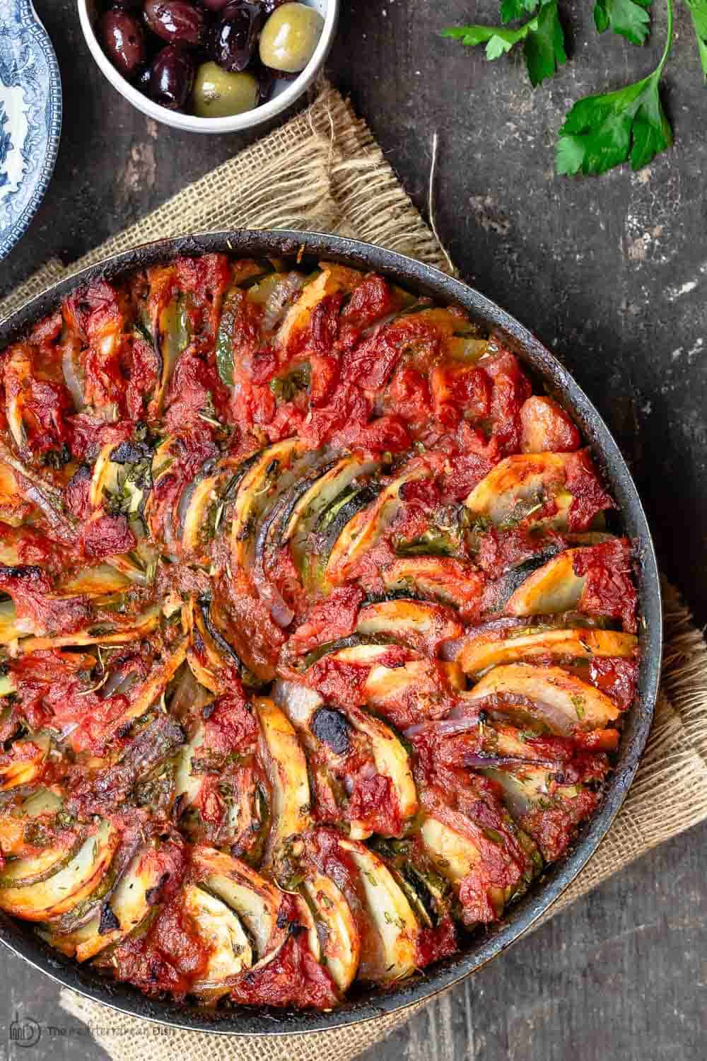 Overhead shot of Briam from The Mediterranean dish with zucchini, potatoes and onion covered in tomato sauce and baked.