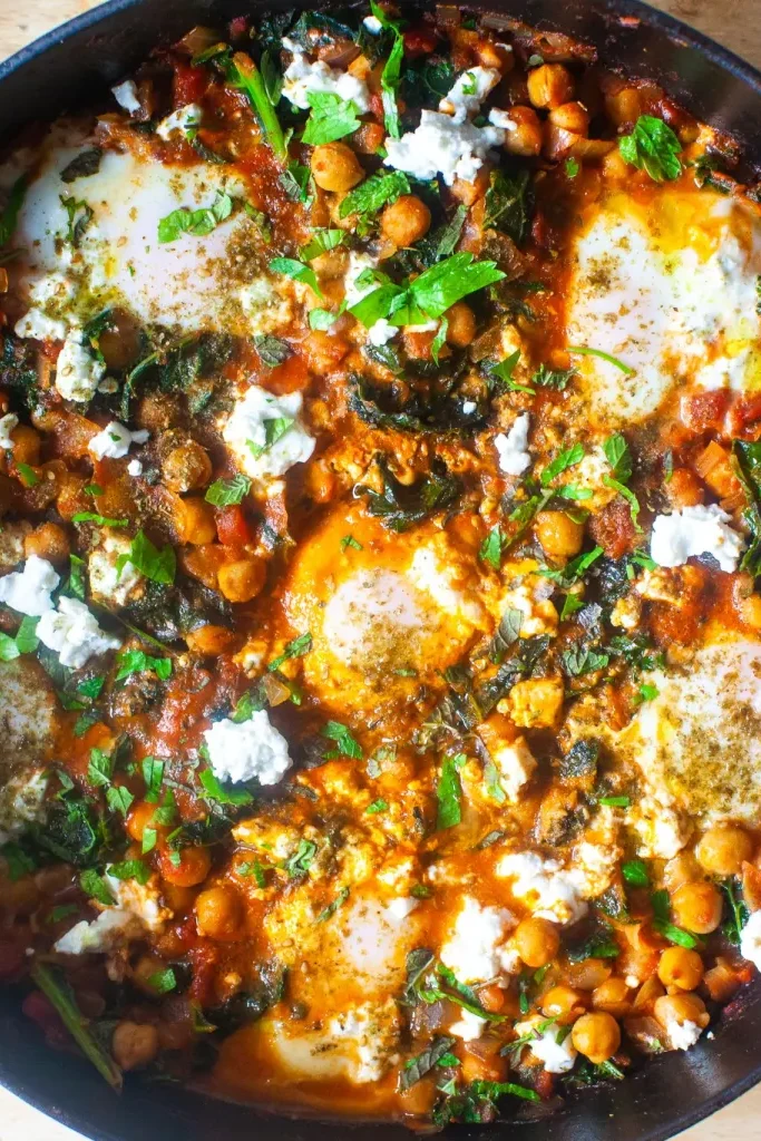 Overhead shot of shakshuka with eggs, chickpeas and kale in a pan.