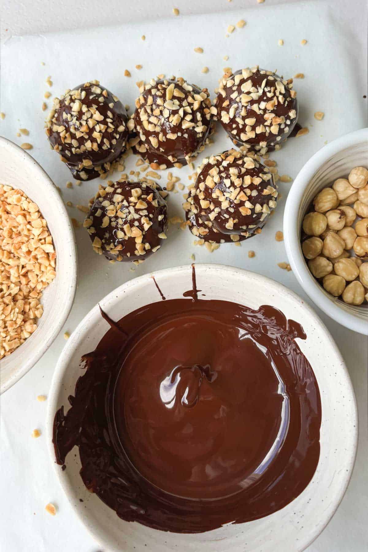 Overhead shot of Ferrero Rocher Protein Truffles above a bowl of melted chocolate, and next to bowls of whole and chopped hazelnuts.
