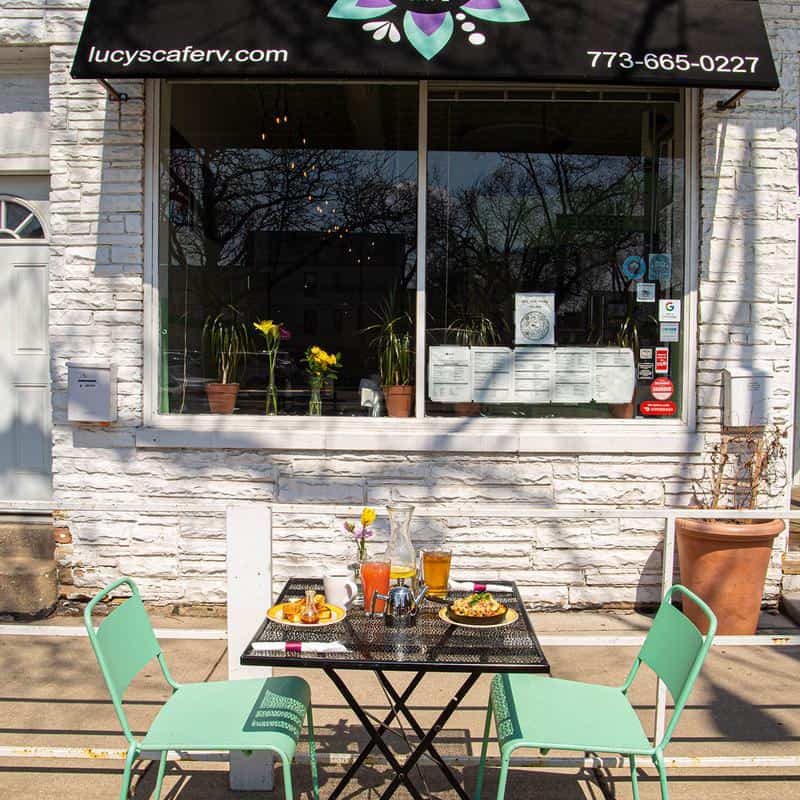 View of the outside of Lucy's cafe featuring a table set with breakfast dishes, with two mint-green chairs.