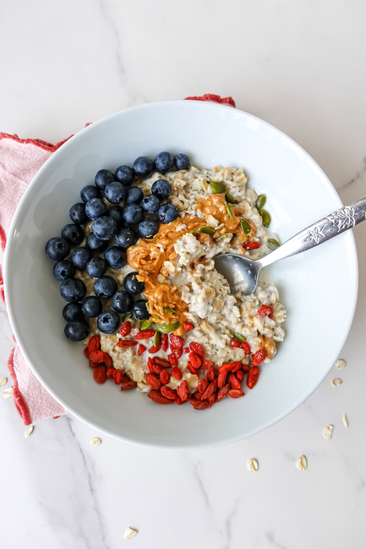 Overhead shot of almond milk porridge in a white bowl, topped with peanut butter, blueberries, goji berries and pumpkin seeds.