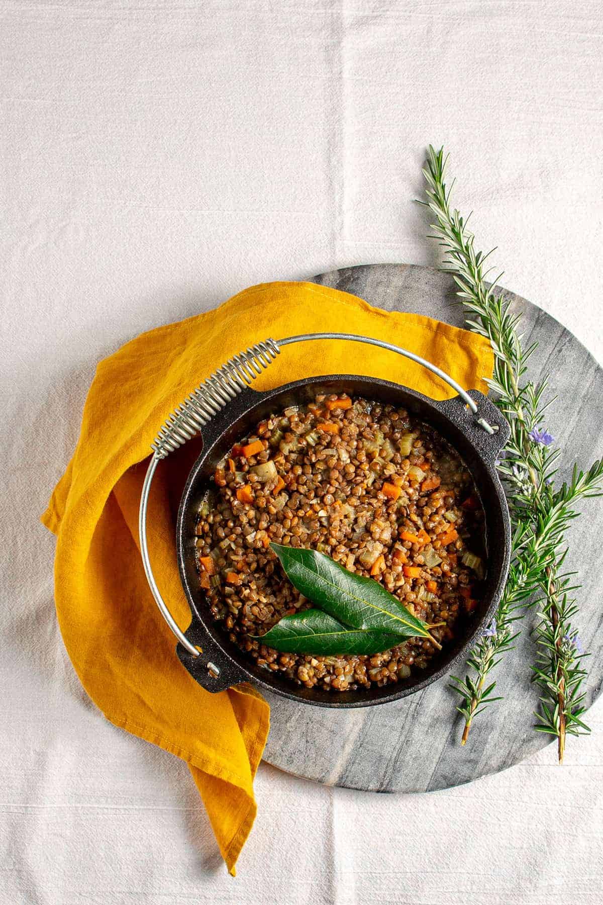 Overhead shot of a small pot of French lentils on a grey stone board, next to rosemary sprigs.