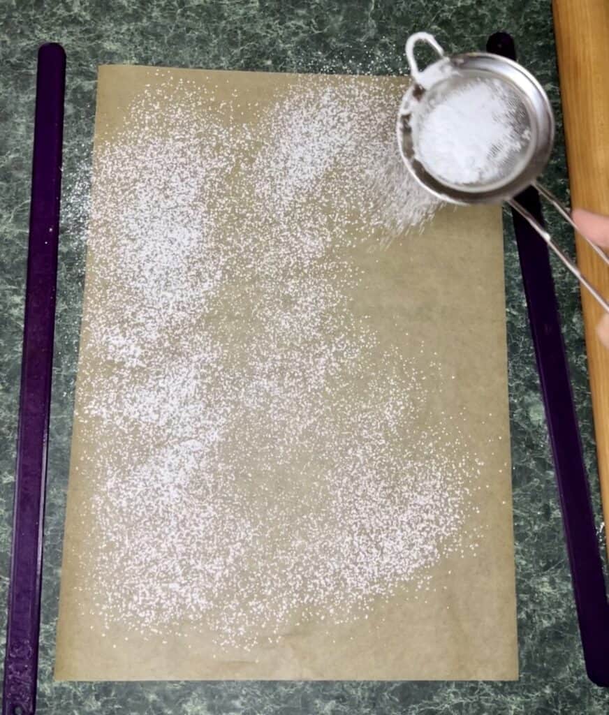 Powdered sugar being sprinkled on parchment paper with a small sifter before rolling the dough 