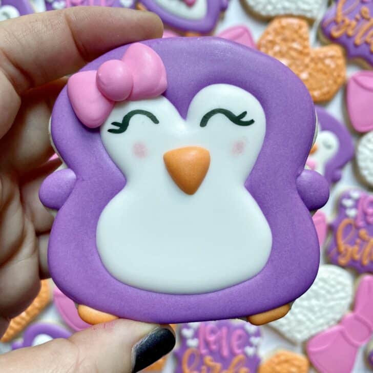 girl penguin cookie decorated with purple, pink, white and orange royal icing