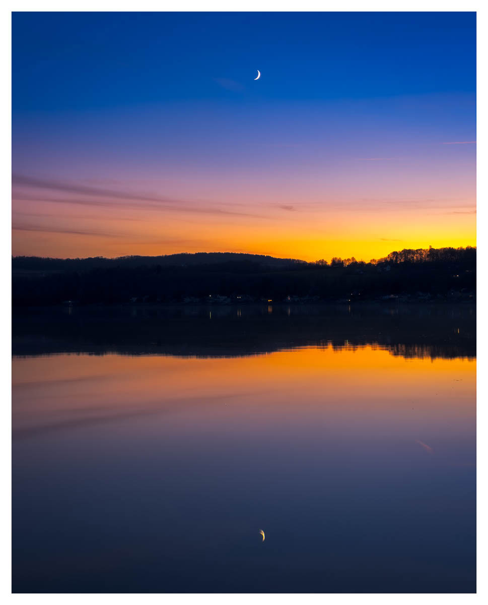 Reflections of the sunset and moon setting over Otisco Lake during blue hour.