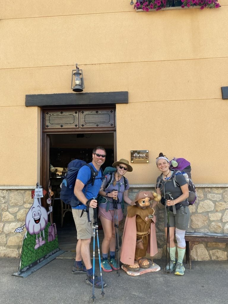 Three hikers smiling outside of a building with a Camino pilgrim statue