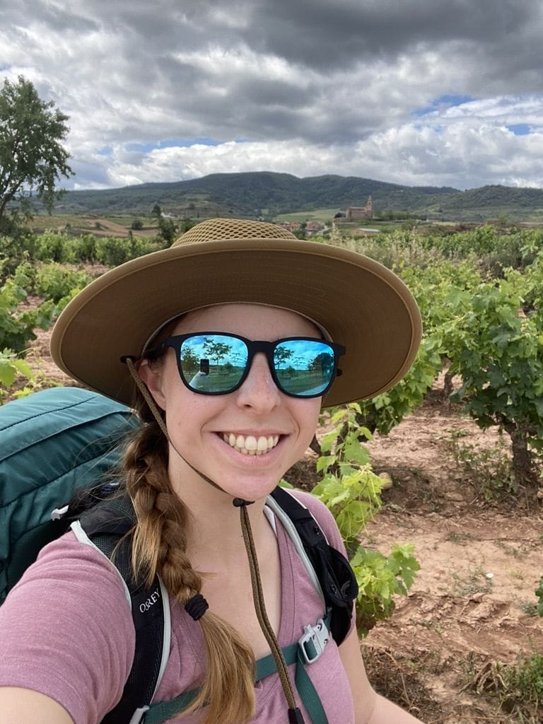 Woman wearing sunglasses and a hat with a backpack smiling with vineyards in the background