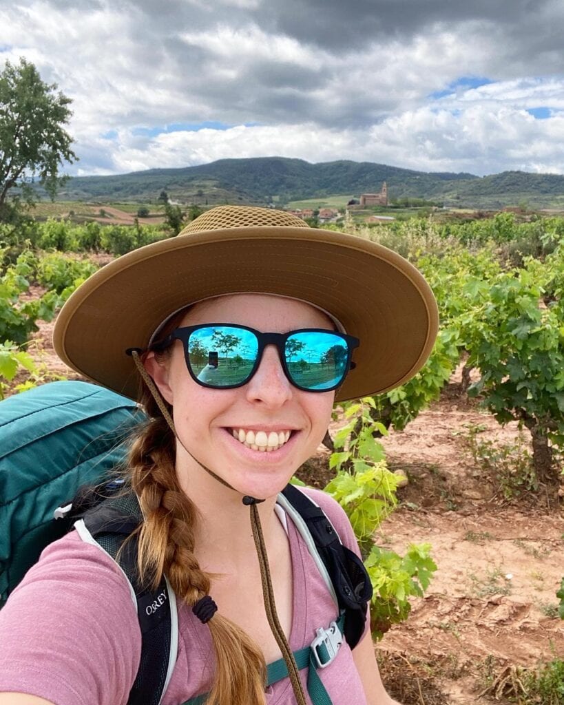 A woman wearing a hat and sunglasses smiling with a small mountain and vineyard in the background along the Camino Francés