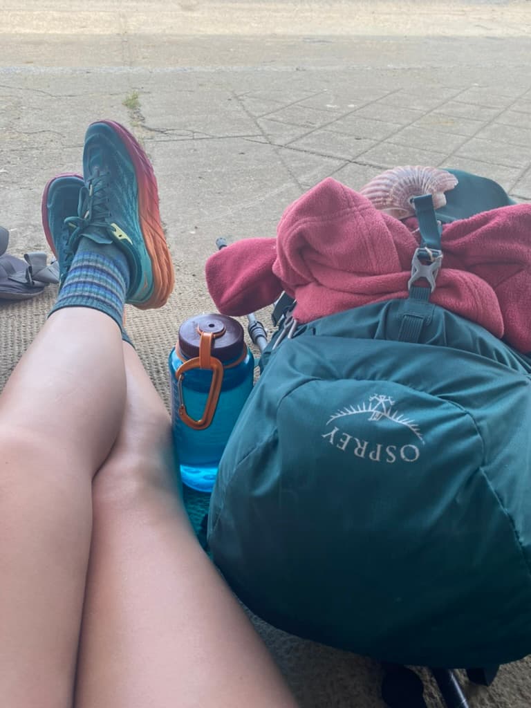 Person's legs crossed sitting on the ground next to a full hiking pack