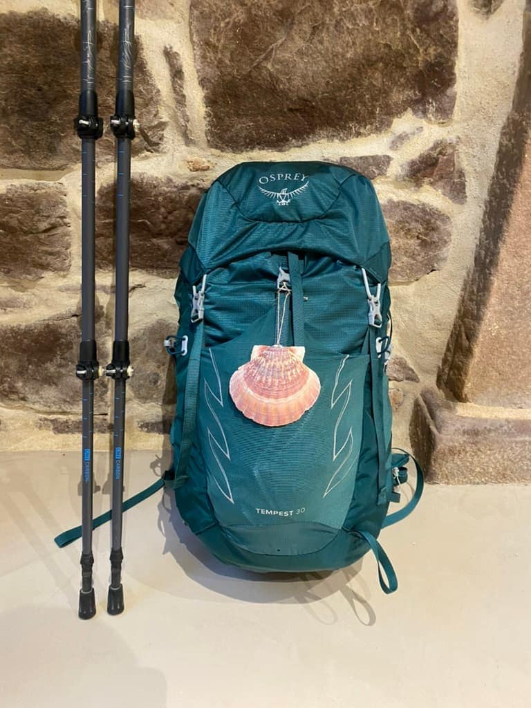 Green hiking backpack with a shell hanging from it and a pair of hiking poles next to it